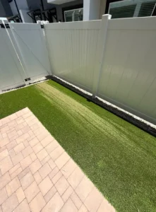 Artificial turf with a long heat-damaged strip caused by sunlight reflecting from a shiny white gate and fence